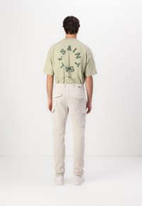 Lys grønn t-skjorte med korte ermer og rund logo på baksiden, kombinert med lyse beige cargo bukser med sidelommer og elastisk liv.