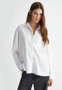 Hvid skjorte med små blå prikker, lange ærmer, en brystlomme og en let afslappet pasform, parret med mørke jeans.