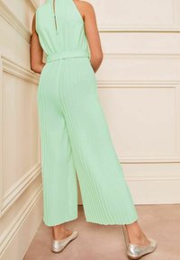 Zielono-miętowy kombinezon z plisami, na halterze, z paskiem w talii i szerokimi nogawkami. Srebrne płaskie buty dopełniają stylizację.