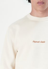 Modellen har en lys beige sweatshirt på, som har "Originals Studio" broderet i brunt på brystet, mod enplain hvid baggrund.