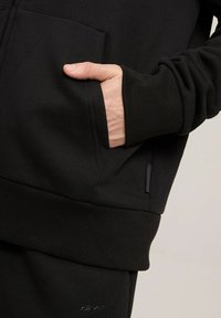 Czarna hoodie z kangurzym kieszeniem, prążkowanymi mankietami i dołem. Miękka tekstura z subtelnym logo marki na dolnej części.