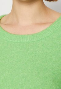 Gros plan sur le cou et la clavicule d'une personne portant un pull en maille douce vert clair avec un col rond.