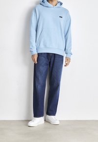 Felpa con cappuccio azzurro chiaro, dotata di tasca frontale e piccolo logo, abbinata a jeans blu scuri a vestibilità ampia e sneakers bianche.