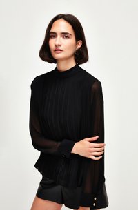 Blusa nera plissettata con maniche lunghe trasparenti, colletto alto e polsini con bottoni dorati. Abbinata a pantaloni corti neri. Tessuto morbido e leggero.