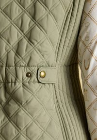 Nahaufnahme einer hellolivgrünen gesteppte Jacke mit diamantförmigen Steppmuster, einem Messing-Druckknopfdetail und einem Teil eines gemusterten cremefarbenen Ärmels.