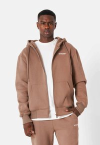Felpa zip-up marrone realizzata in tessuto morbido, con tasca frontale, cappuccio regolabile con cordini e logo bianco sul lato sinistro del petto. Abbinata a una maglietta bianca.
