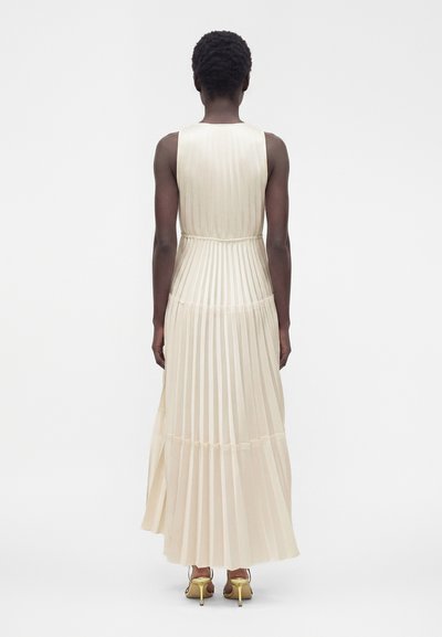 Personne portant une robe maxi plissée sans manches crème et des sandales à talons dorés, debout, dos tourné, sur un fond blanc uni.