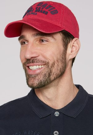 Red cotton cap with embroidered text, featuring a curved brim. The individual has short hair and a beard, wearing a dark polo shirt.