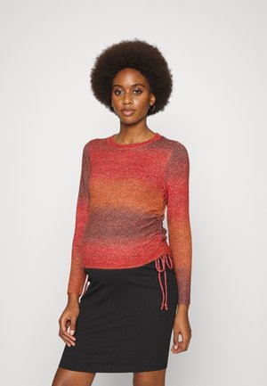 Suéter com gradiente vermelho e laranja, de mangas longas, com textura canelada e detalhes de cordão nas laterais. Combinado com uma saia canelada preta.