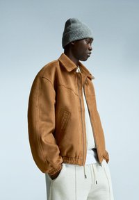 Veste en cuir suédé marron avec un col, une fermeture éclair à l'avant et des poignets élastiques, portée sur un sweatshirt gris clair. Accompagnée d'un bonnet en tricot gris.