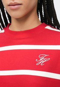 Sweatshirt en coton rouge avec des rayures horizontales blanches, col rond et logo brodé sur la poitrine. Texture lisse.