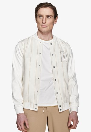 Mężczyzna w kremowej kurtce varsity z napami, białej koszuli i beżowych spodniach, patrzący lekko w lewo.