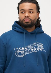 Modrá bavlněná mikina s kapucí s grafickým potiskem a vrstveným textem "JACK & JONES 1990" v bílé barvě. Má stahovací kapuci a prostorný střih.