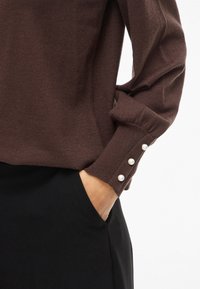 Rudas megztinis ilgomis rankovėmis, su trimis perlamutriniais mygtukais rankogalyje. Audinys atrodo minkštas ir kostiumuotas, derinamas su juodu apatiniu drabužiu.