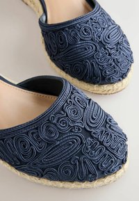 Chaussures espadrilles bleu marine avec une broderie tourbillonnante en relief détaillée et des semelles en jute tressé, présentées en gros plan sur un fond clair.