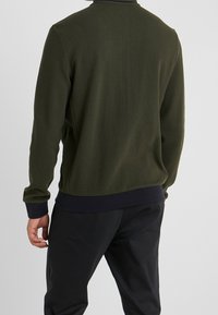 Dunkelgrünes strukturiertes Sweatshirt mit langen Ärmeln und einem gerippten marineblauen Saum. Auffällige Merkmale sind ein Stehkragen und eine lockere Passform.