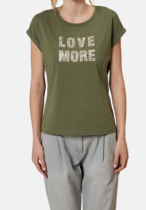 Kobieta nosząca oliwkowo zielony T-shirt z podwiniętymi rękawami i białym nadrukiem w cekiny "LOVE MORE", w połączeniu z luźnymi jasnoszarymi spodniami.