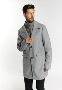 Grau melierter Wollmantel mit Reverskragen, zwei Fronttaschen und zwei Knöpfen. Darunter befindet sich ein graues, gestepptes Innenfutter.