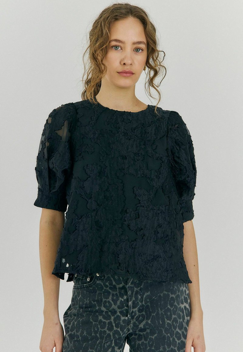 Blusa corta de manga corta texturizada negra con un patrón floral y acentos transparentes. Combinada con unos vaqueros oscuros de estampado de leopardo.