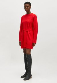 Rotes gestricktes Kleid mit Taillengurt, lange Ärmel und lässige Passform. Getragen mit eng anliegenden schwarzen kniehohen Stiefeln.