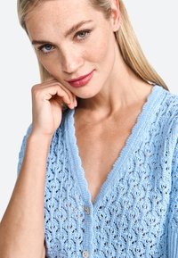 Cardigan lavorato a maglia azzurro chiaro con un delicato motivo in pizzo, scollo ondulato e bottoni decorativi. Il tessuto testurizzato aggiunge interesse visivo.
