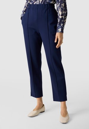 Kobieta ubraną w granatowe, proste spodnie o długości do kostek z przednimi kantami, elastyczną talią, bocznymi kieszeniami, wzorzystą bluzkę i kremowe płaskie buty.