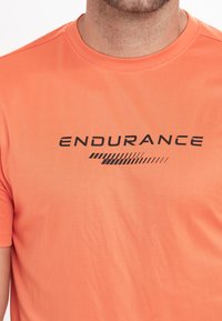 Oranssi urheilu t-paita, joka on valmistettu kevyestä materiaalista ja jossa on musta "ENDURANCE" -logo ja graafiset elementit edessä.