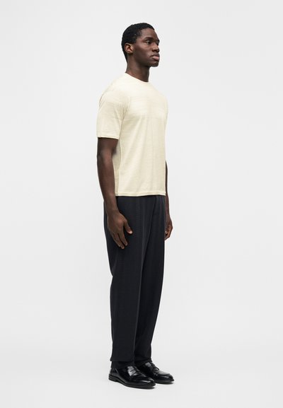 Ανοιχτό μπεζ κοντομάνικο μπλουζάκι με διακριτικό υφασμάτινο μοτίβο, συνδυασμένο με άνετο μαύρο παντελόνι με ψαροκόκαλο και μαύρα παπούτσια.