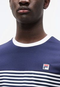 Σκούρο μπλε μπλούζα με κοντά μανίκια και λευκές ρίγες, με στρογγυλή λαιμόκοψη και λογότυπο FILA στο αριστερό μέρος του στήθους. Υφασμάτινη υφή λεία.