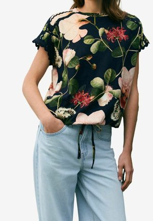 Kvinde iført en marineblå kortærmet top med store pink og røde blomster, kombineret med lyseblå højttaljede jeans, den ene hånd i lommen.