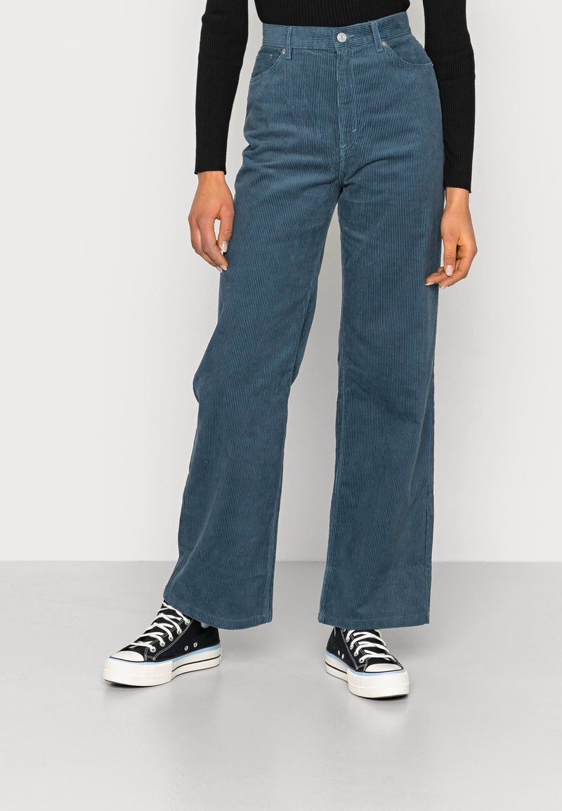 Bredvids corduroybyxor i skifferblått, med vertikala ribbade mönster, hög midja och framfickor, tillsammans med svarta sneakers.