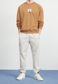 Person som bär en beige sweatshirt och krämfärgade cargobyxor med händerna i fickorna, stående på en blå matta mot en vit vägg.