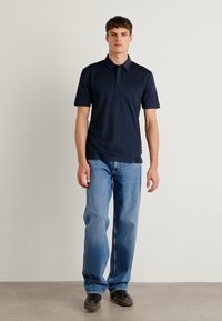 Camisa polo azul-marinho de manga curta com gola, combinada com jeans azul claro e calçado preto, posando contra um fundo neutro.