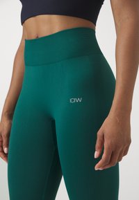 High-waisted green leggings made of stretchy fabric, featuring a textured waistband and a small white logo on the thigh.