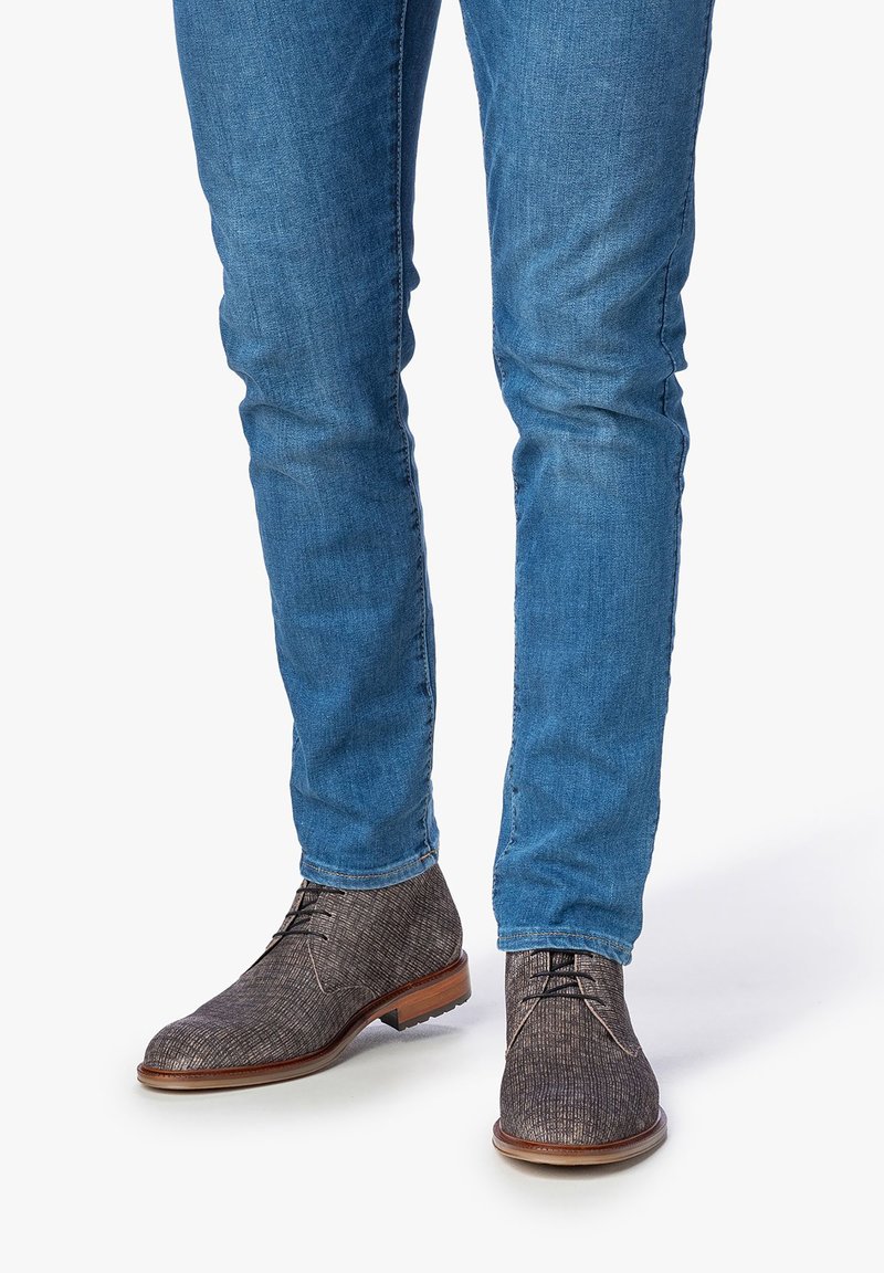 Stivali alla caviglia marroni con texture e un sottile motivo, abbinati a jeans blu attillati, con suola in pelle liscia e lacci.
