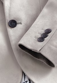 Hellgrauer Wildlederblazer mit Reverskragen, schwarzen Knöpfen und dezenten Steppnähten an den Manschetten. Weiche Textur, maßgeschneiderte Passform.