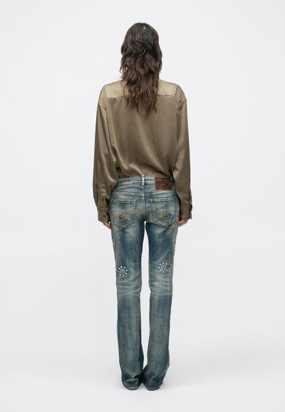 Osoba s dlhými vlnitými vlasmi, oblečená v voľnej hnedej saténovej košeli a zdobených modrých džínsoch, stojaca chrbtom k fotoaparátu na neutrálnom pozadí.