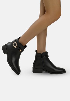 Botines negros de tobillo hechos de cuero sintético suave, con cremallera lateral, una correa ajustable con un acento metálico redondo y un tacón bajo.