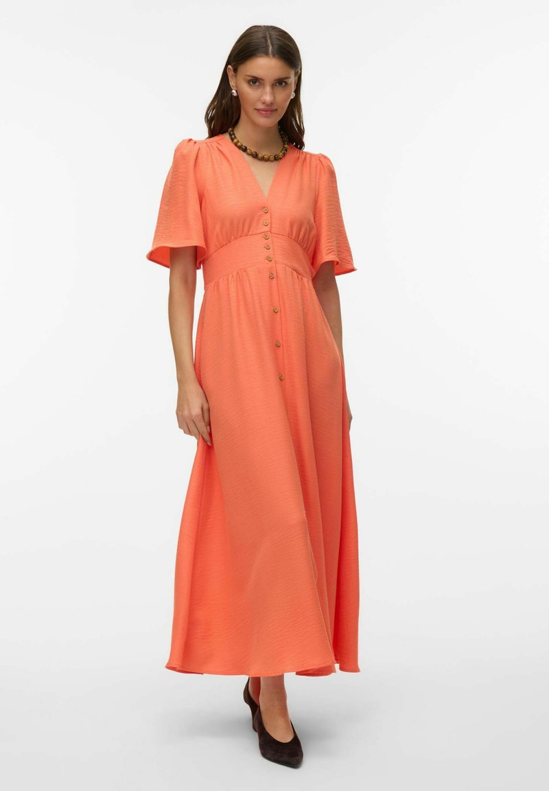 Femme portant une robe longue corail orange avec des manches courtes à volants, boutonnée sur le devant, associée à des talons noirs et un collier de perles.