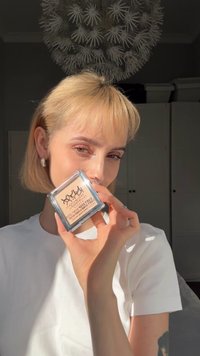 Transluzente quadratische Kompaktverpackung mit beigem Puderprodukt, das ein Logo auf der Oberseite zeigt. Model hält es vor einem hellen Hintergrund.