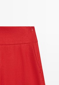 Červená sukně s vysokým pasem, skrytým bočním zipem a hladkou texturou látky, zobrazena na jednoduchém bílém pozadí.