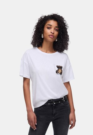 Donna con capelli ricci che indossa una maglietta bianca con un piccolo disegno di orsetto e palloncino, cintura nera e jeans scuri su uno sfondo neutro.