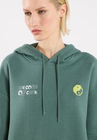 Felpa verde realizzata in morbido tessuto, con grafiche bianche e gialle, compresi testo e simbolo yin-yang sulla parte anteriore.