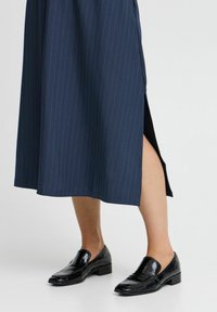 Jupe midi bleu marine à rayures avec fente latérale, en tissu lisse. Associée à des mocassins noirs en cuir verni avec un petit talon et un bout pointu.