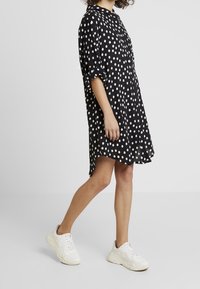 Schwarzes Kurzarmkleid mit weißen Punkten; lockere Passform, knielang und abgerundeter Saum. Kombiniert mit weißen Turnschuhen.