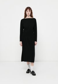 Črna dolgo rokavna obleka z čolnastim izrezom, oprijetim telovnikom in tekočo krilom. Oblečena z črnimi loaferji in belimi nogavicami. Minimalističen dizajn.
