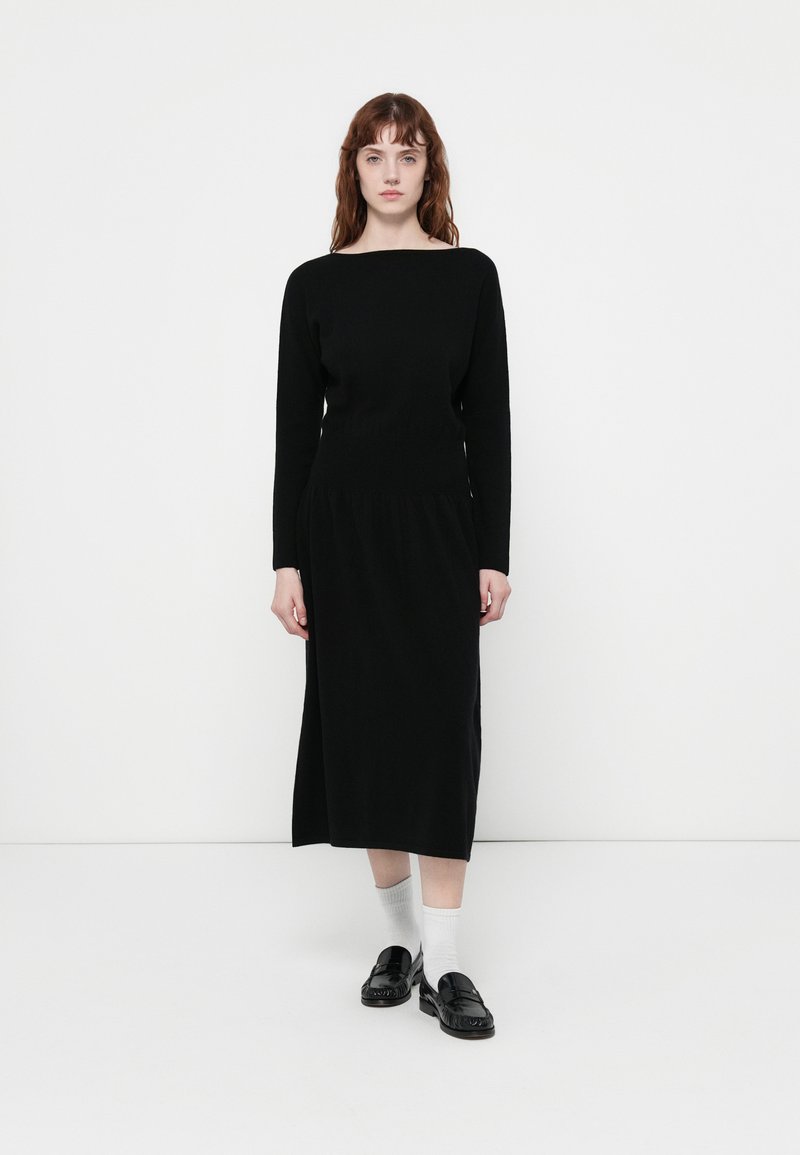 Abito lungo nero con maniche a lunghe, scollatura a barca, corsetto aderente e gonna fluente. Indossato con mocassini neri e calzini bianchi. Design minimalista.