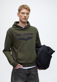 Oliwkowa bluza z kapturem z przednią kieszenią i wyszywanym logo "PME LEGEND AMERICAN CLASSIC". Model trzyma czarną torbę. Wyblakłe czarne jeansy.