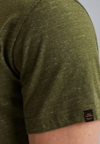 Olivegrünes Baumwoll-T-Shirt mit strukturiertem Finish. Kurze Ärmel, rundhalsausschnitt, mit einem kleinen gewebten Etikett am unteren Saum.