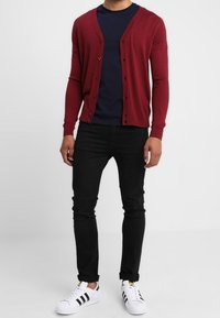 Cárdigan burdeos con un diseño de escote en V, usado sobre una camisa azul marino, combinado con jeans negros y zapatillas blancas con rayas negras.